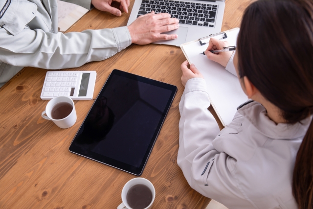 木のテーブルで打ち合わせする2人。ノートPCとクリップボードを使っている